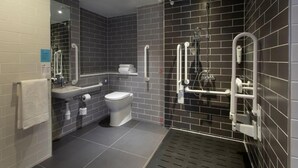 Un baño moderno con inodoro, lavamanos y zona de ducha, con azulejos oscuros y barras de apoyo.