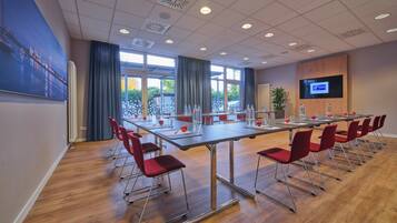A conference room with a long table, red chairs, a large window, and a screen on the wall.