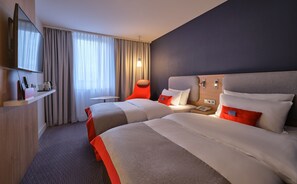 Ein Hotelzimmer mit zwei Betten, einem Fernseher, einem Schreibtisch mit Telefon und einem Fenster mit Vorhängen.