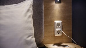 Una cama bien hecha con un almohadón blanco, una mesita de noche con un teléfono inteligente y su cargador, y un enchufe en la pared.