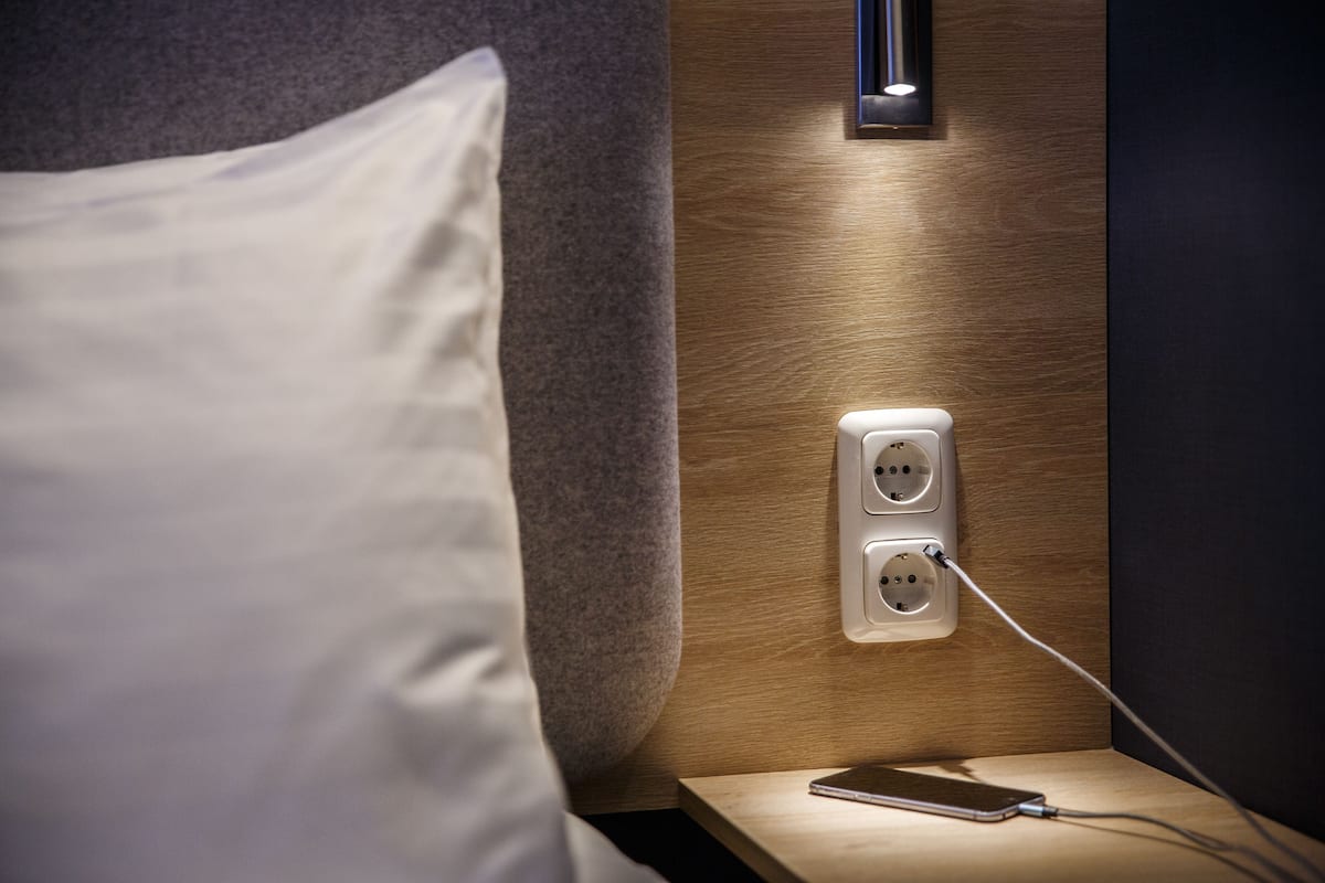Un lit bien fait, avec un oreiller blanc, une table de chevet sur laquelle se trouvent un téléphone intelligent et un chargeur, et une prise de courant fixée au mur.