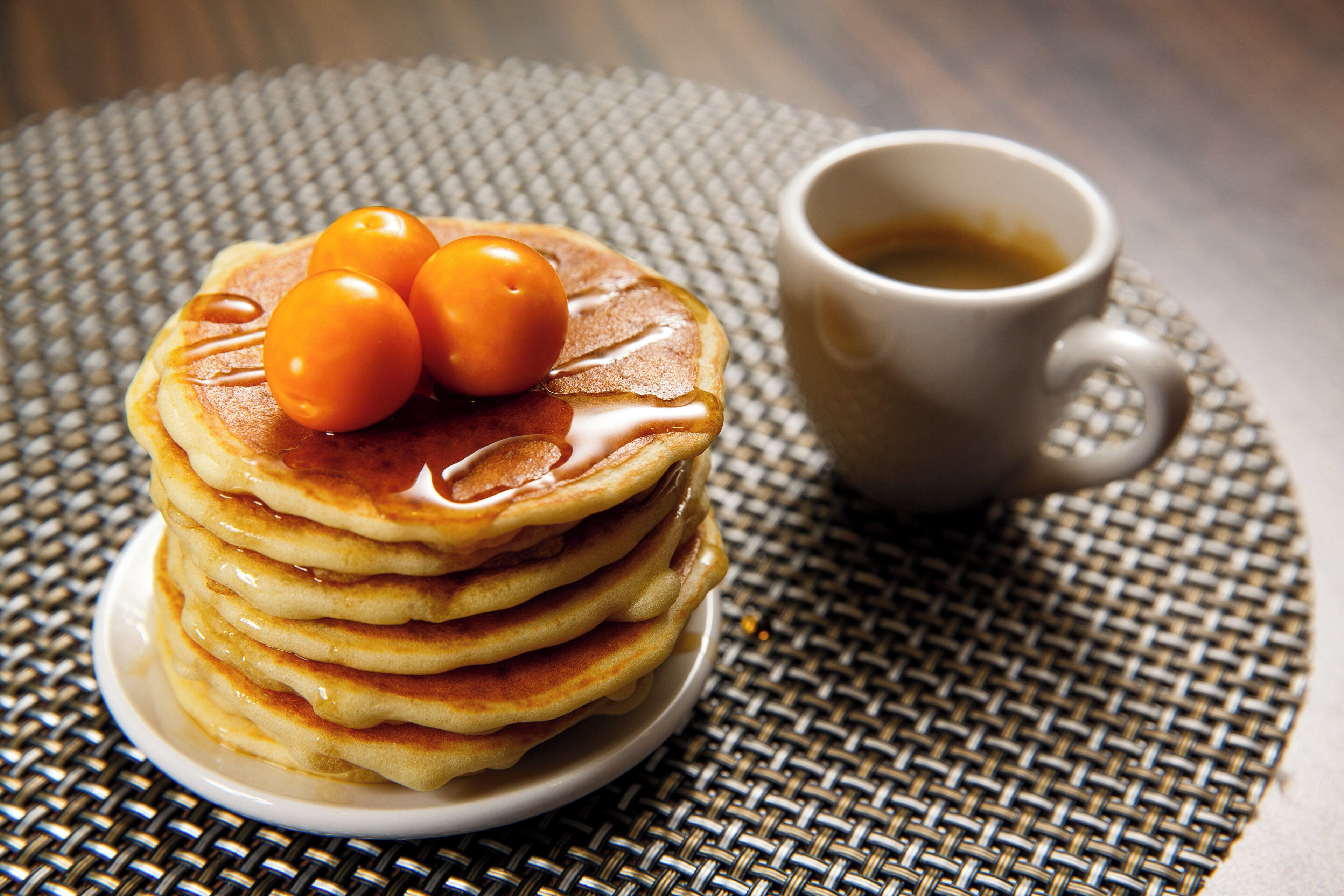 Una pila de tortitas con jarabe y dos huevos encima, sobre un posavasos tejido, junto a una taza de café.