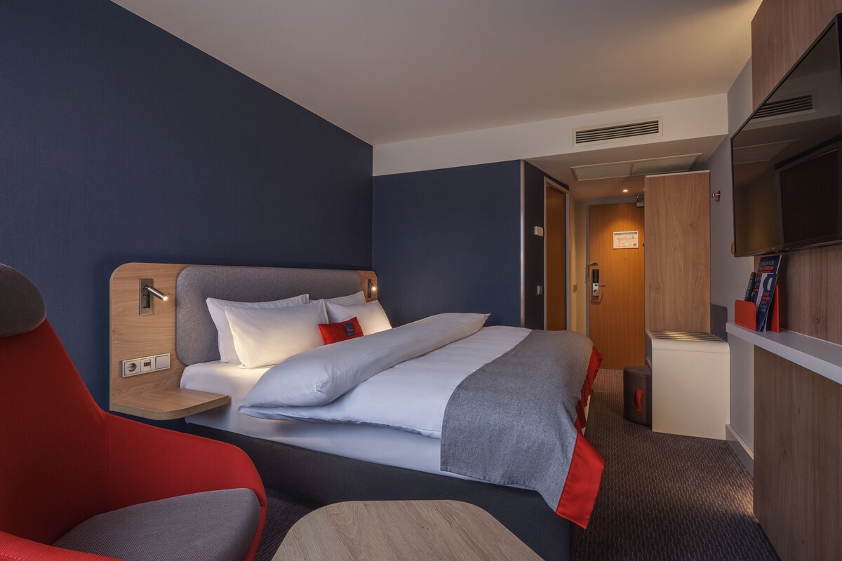 A hotel room with a bed, a TV mounted on the wall, a desk with a lamp, and a red chair.