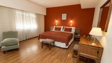 A hotel room with a wooden floor, a bed with a red bedspread, a green armchair, a nightstand, a bedside table with a lamp, and a large mirror.