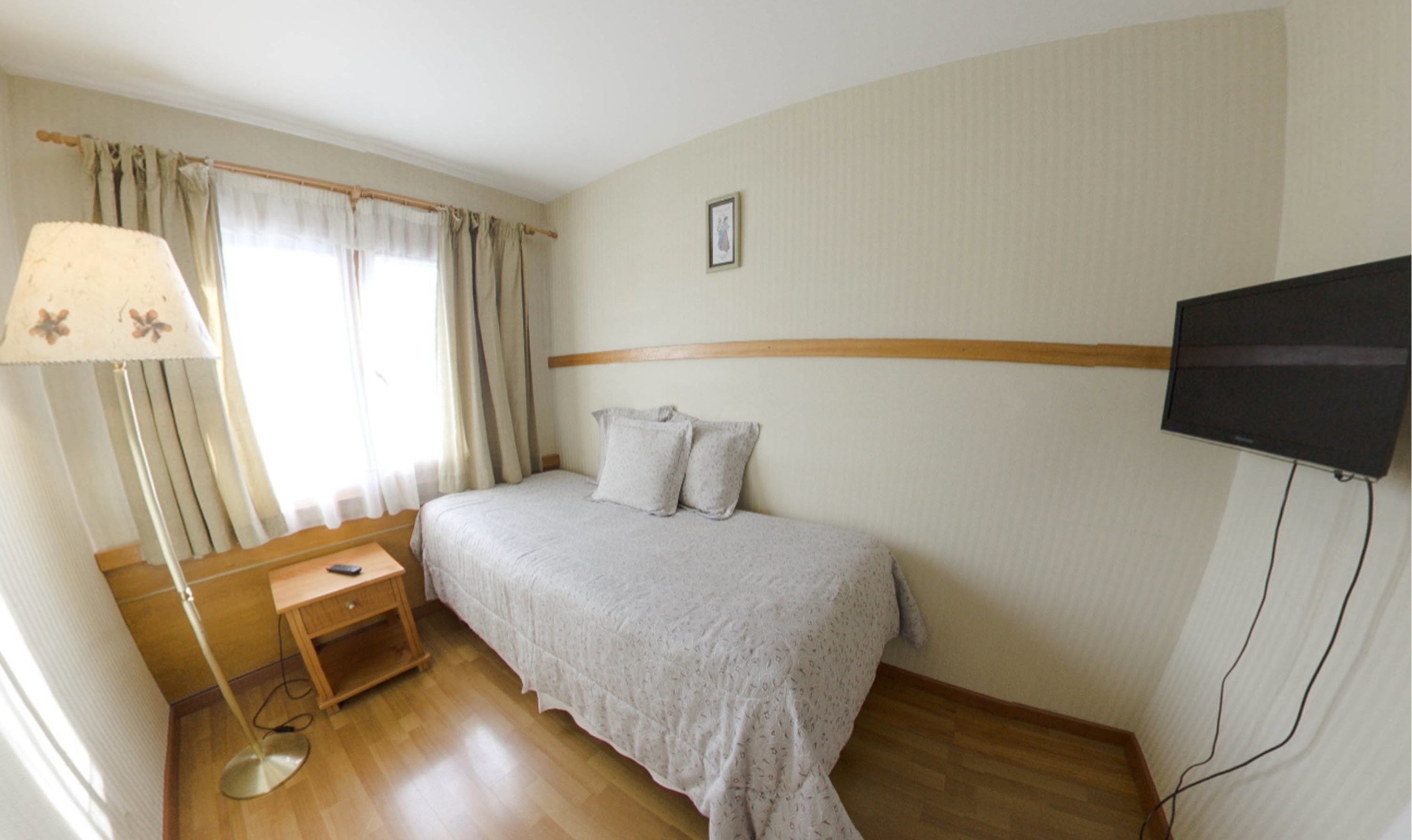 Un dormitorio con cama, mesita de noche, lámpara, televisión y ventana con cortinas.