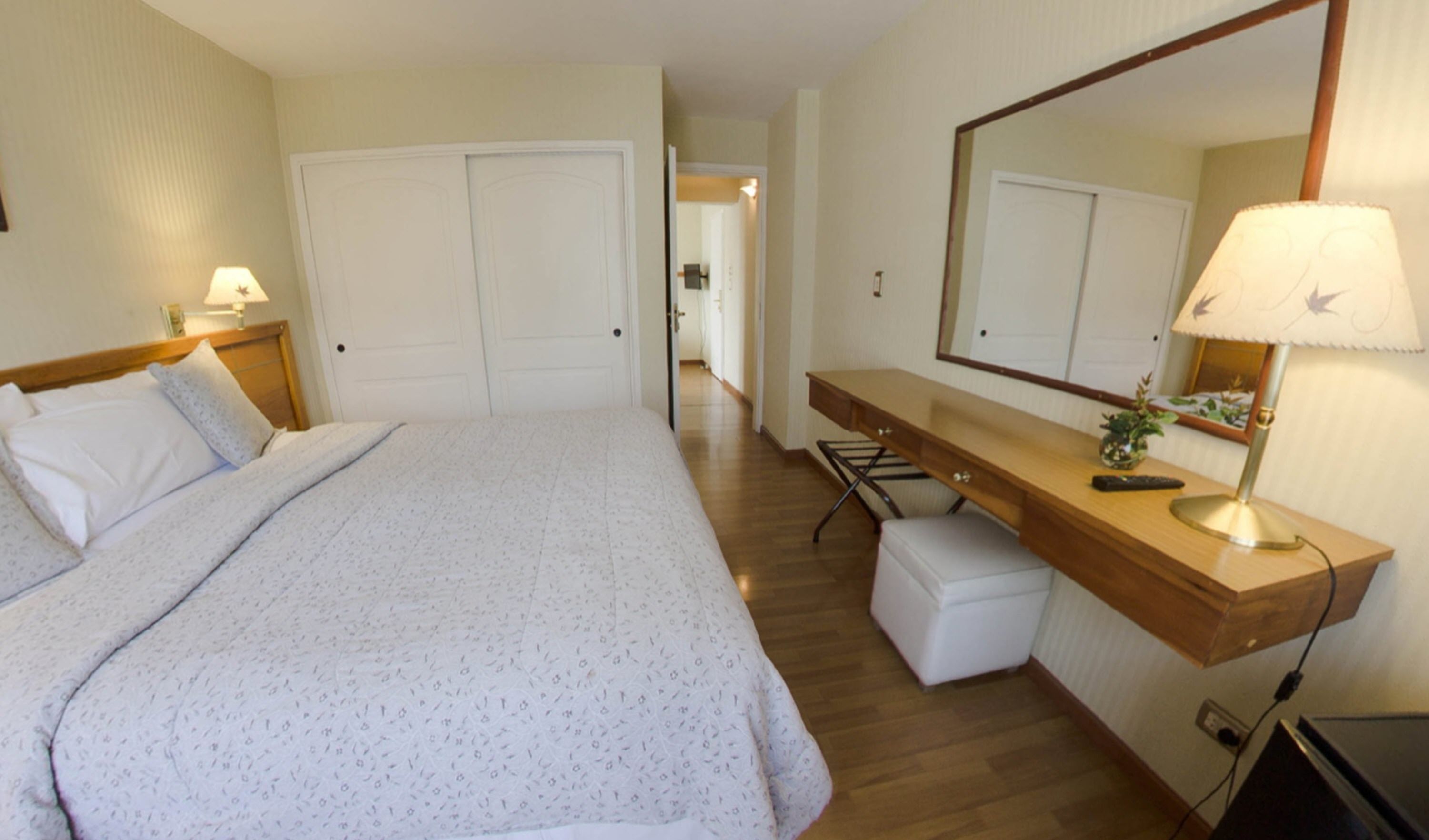 A bedroom with a bed, a desk, a chair, a mirror, and a lamp.