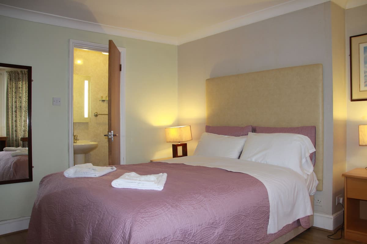 Habitación de hotel con cama, mesita de noche, lámpara, espejo y baño visible a través de una puerta abierta.