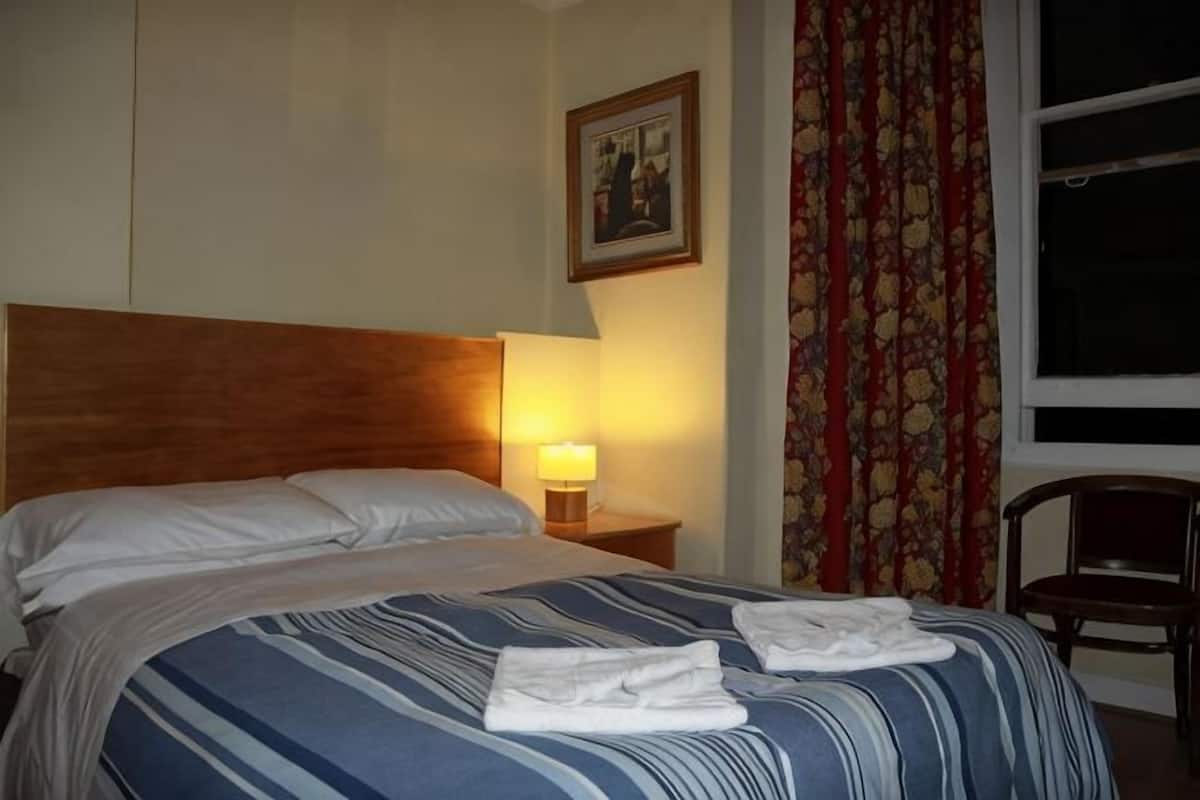Una habitación de hotel con una cama, una mesita de noche con lámpara, una silla y una ventana con cortinas.