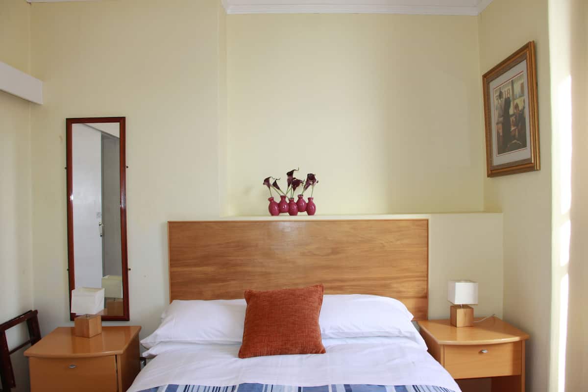 Habitación de hotel con cabecera de madera, cama con sábanas blancas, almohada decorativa roja, mesita de noche con lámpara y un florero sobre la mesita.
