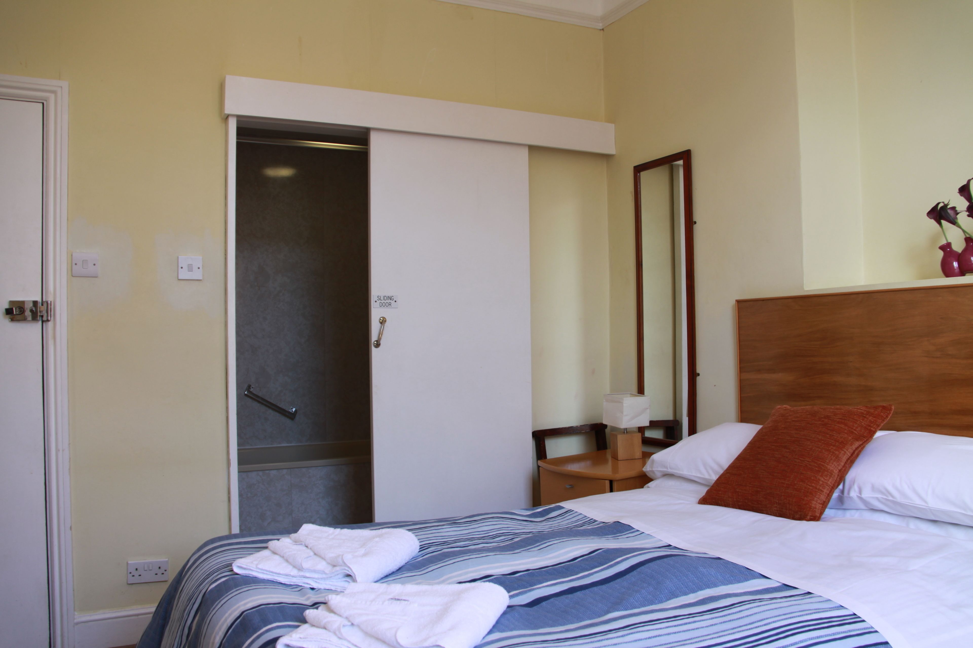 A hotel room with a bed, a wooden headboard, a nightstand, a mirror, and a sliding door to a bathroom.