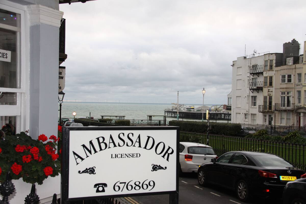 Señal que indica 'Ambassador Licensed' con un número de teléfono, cerca de una zona costera con coches aparcados y edificios al fondo.