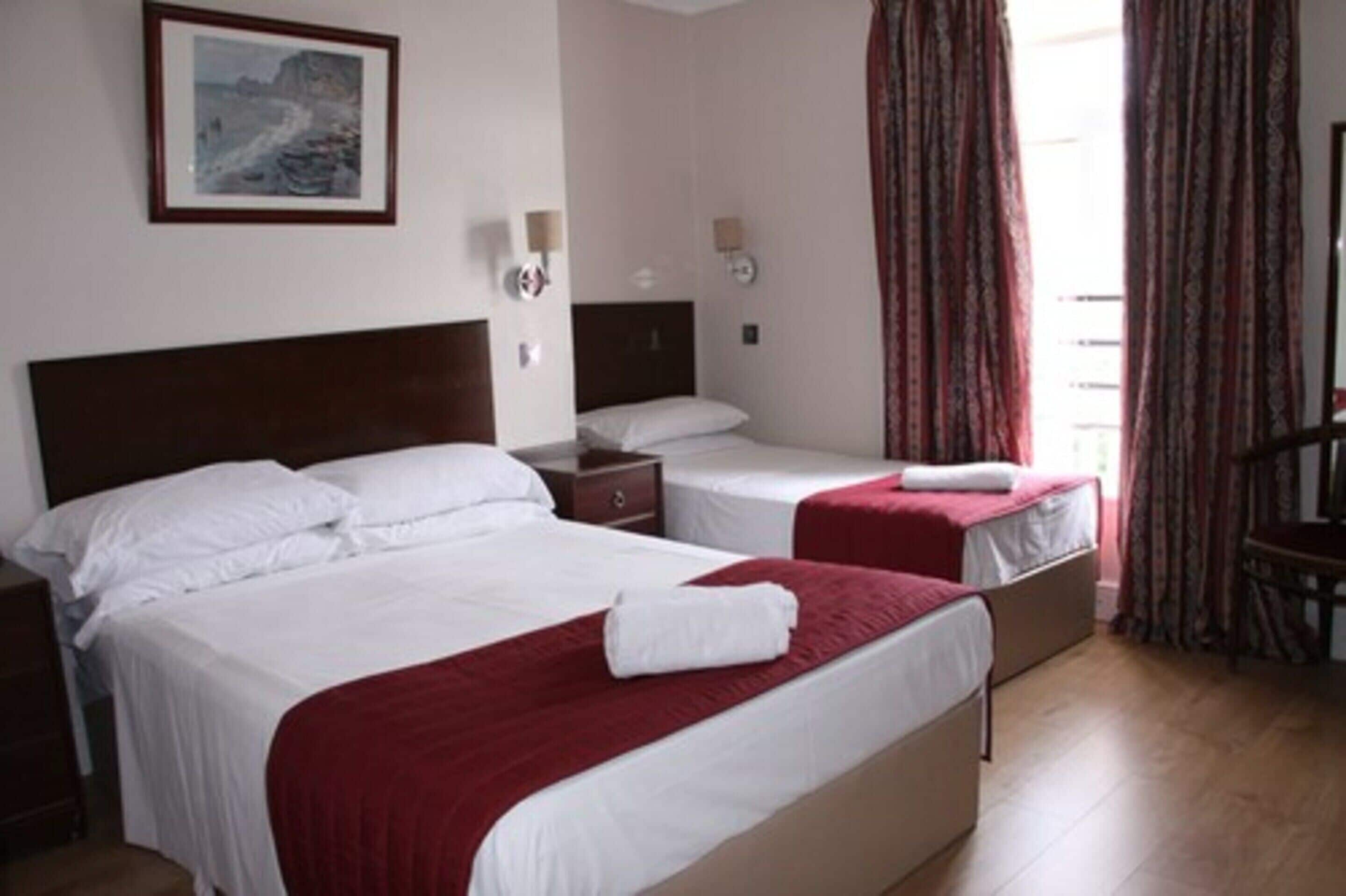 Quarto de hotel com duas camas, uma mesa de cabeceira, uma luminária na parede e uma janela com cortinas.