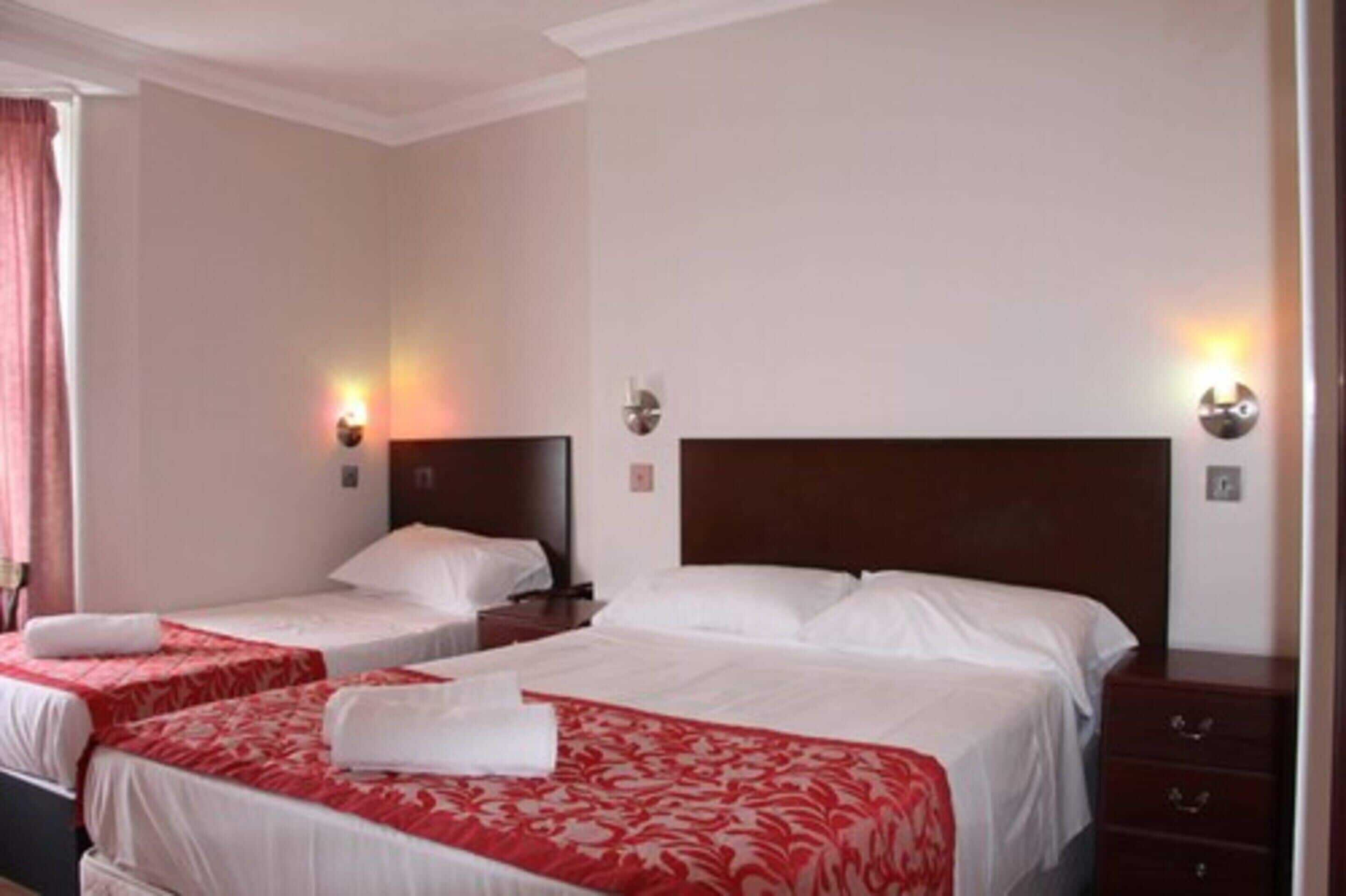 A hotel room with two single beds, each with a red and white bedspread, and a wooden headboard. There are wall-mounted light fixtures on either side of the beds.