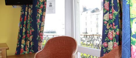 Una habitación con un escritorio de madera, dos sillas anaranjadas, cortinas con motivos florales, un televisor montado en la pared y una ventana con vistas a edificios.