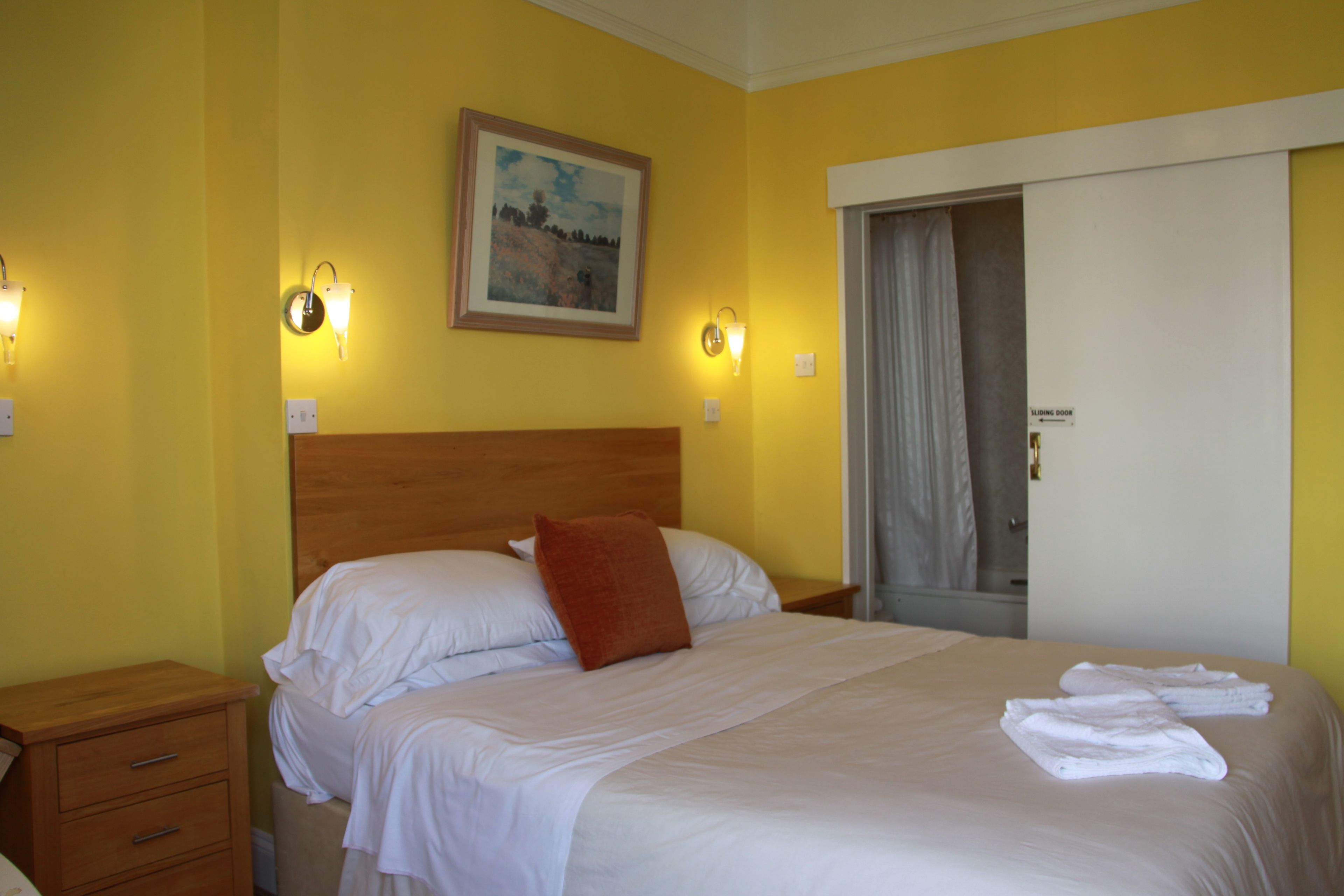 A hotel room with a bed, a nightstand, a wooden headboard, a painting on the wall, and a door with a curtain.