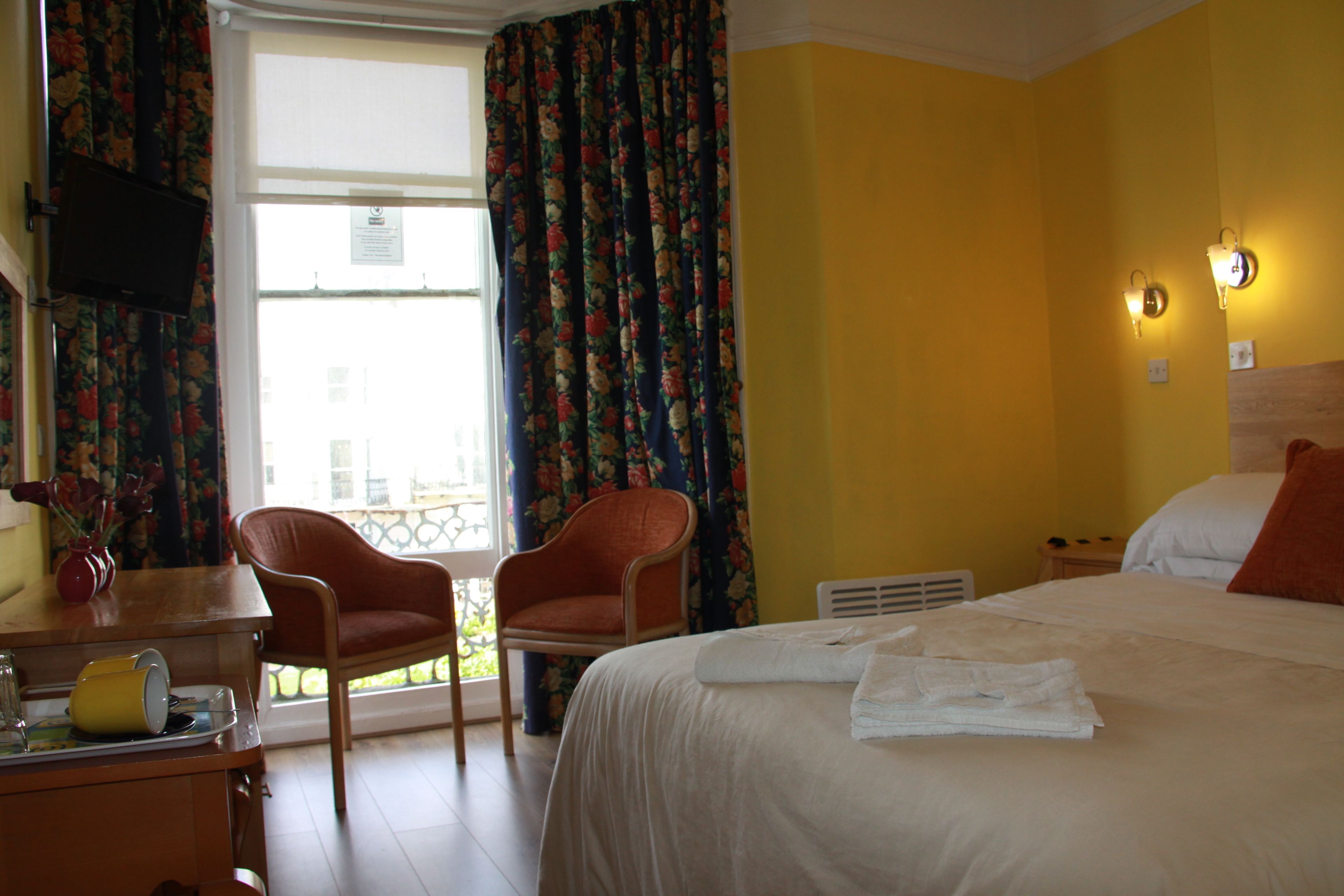 A hotel room with a bed, two chairs, a desk with a telephone, a window with curtains, and a wall-mounted light fixture.