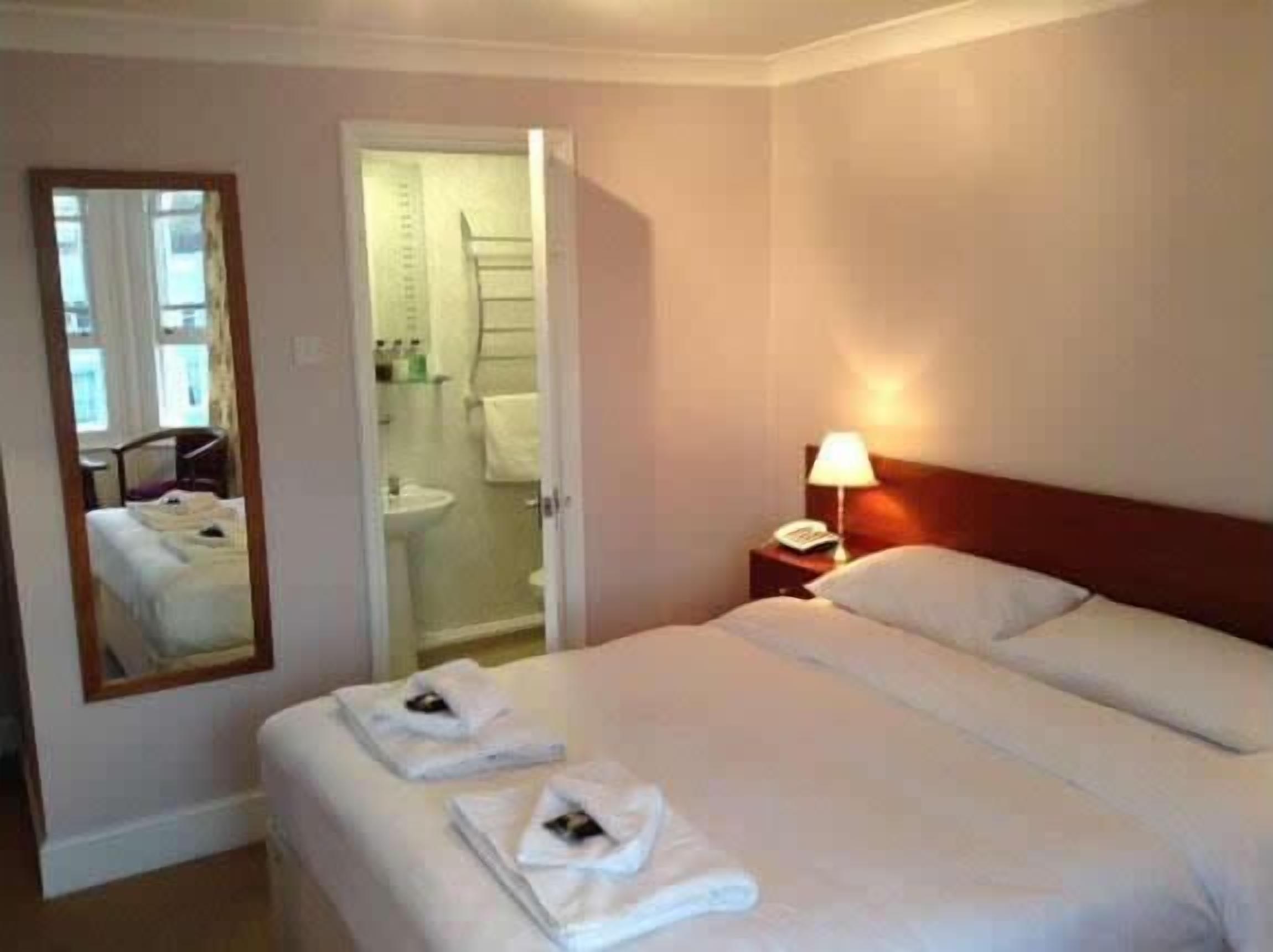 Una habitación de hotel con una cama, una mesita de noche con lámpara, un espejo y un baño visible a través de una puerta abierta.