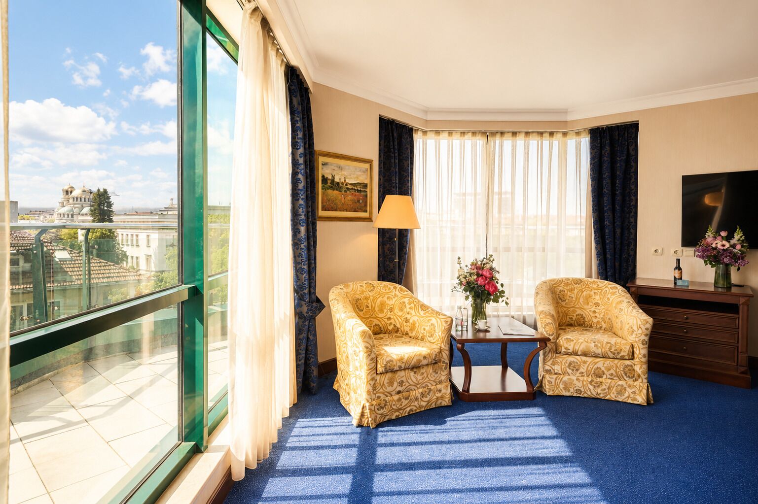 A hotel room with two patterned armchairs, a small table with a vase of flowers, a painting on the wall, and a view of a building through large windows.