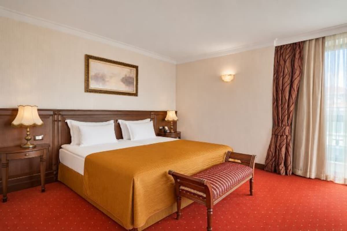 Quarto de hotel com uma cama grande, mesas de cabeceira com luminárias, um banco, um quadro na parede e uma janela com cortinas.