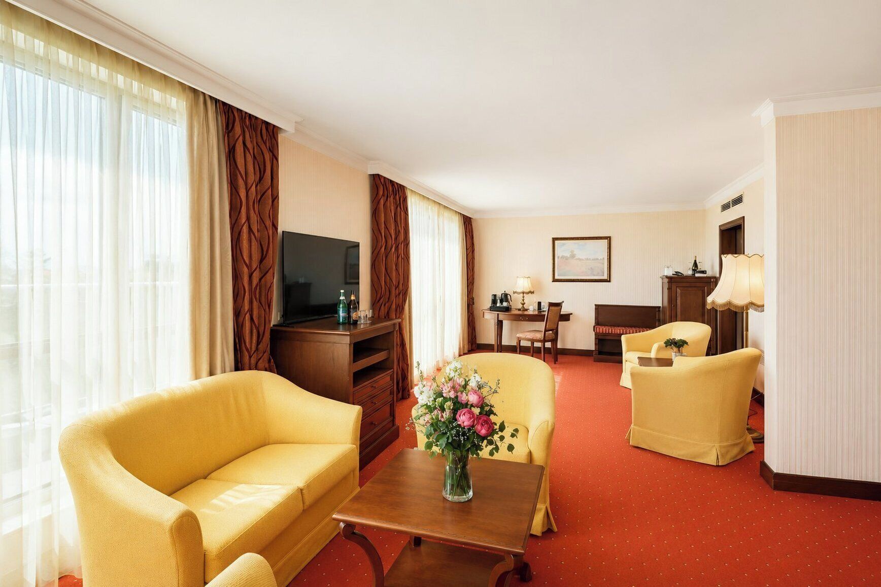 Une chambre d’hôtel comprenant un canapé, un fauteuil, une petite table avec un vase de fleurs, un téléviseur posé sur un meuble en bois, un bureau avec une chaise et une lampe.