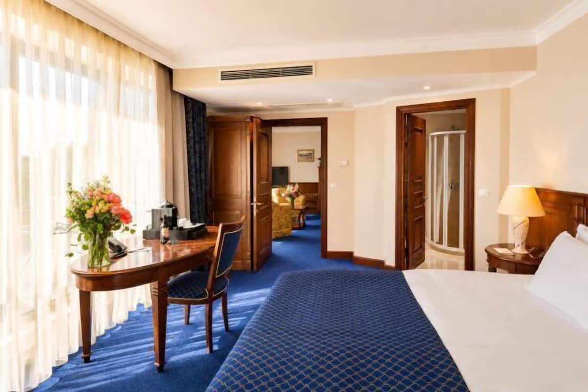 A hotel room with a bed, a desk with a chair, a lamp, and a view of a hallway.