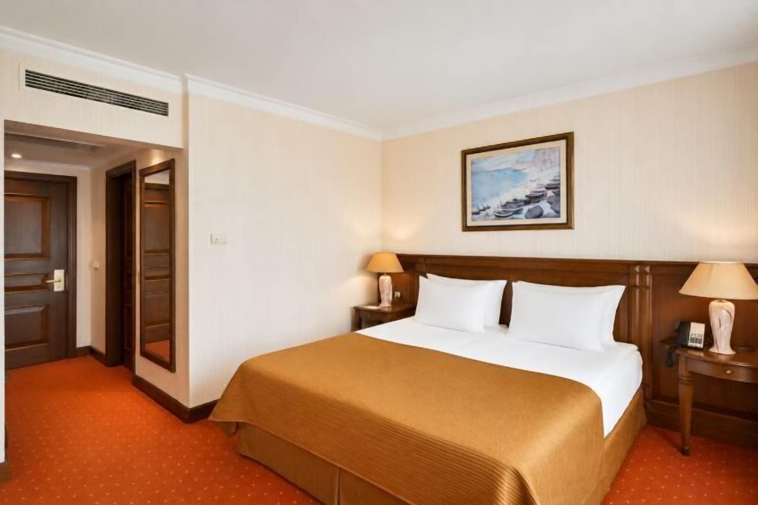 Une chambre d’hôtel avec un grand lit, deux tables de chevet, une commode, un tableau au mur et une porte sur le côté gauche.