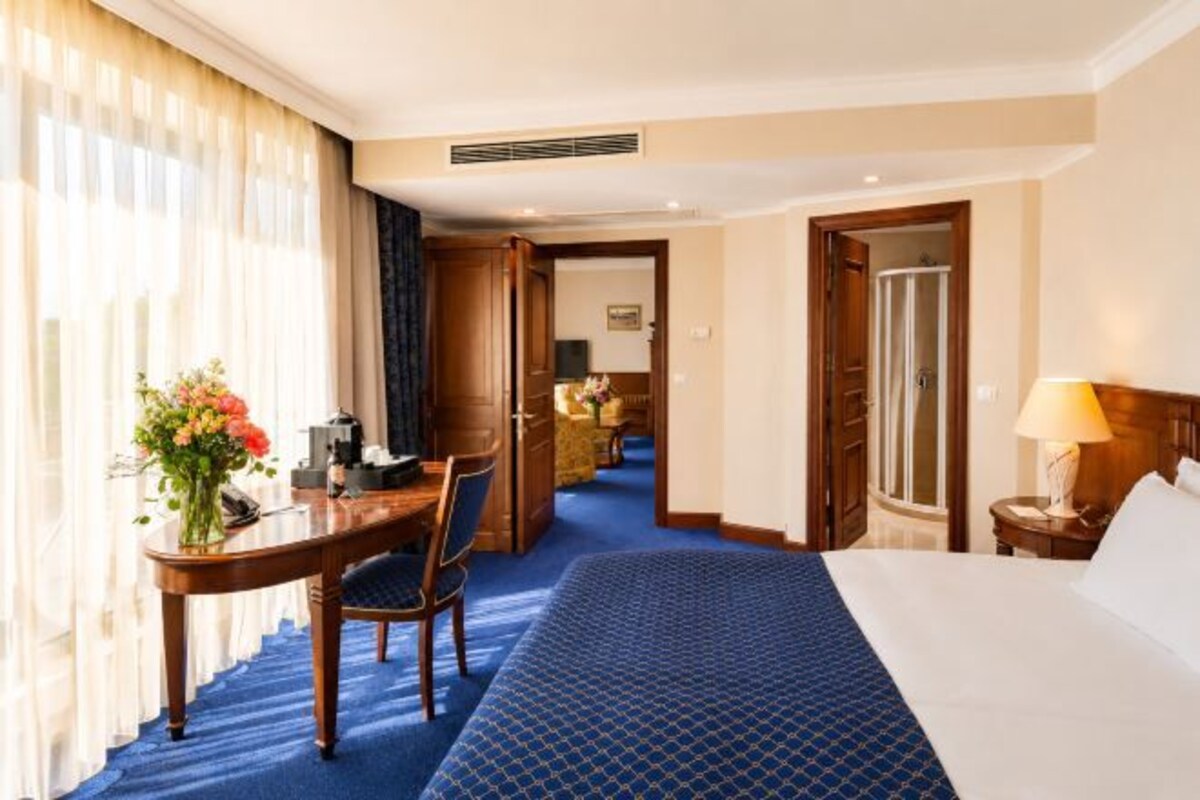 A hotel room with a bed, a desk with a chair, a lamp, and a view of a hallway.