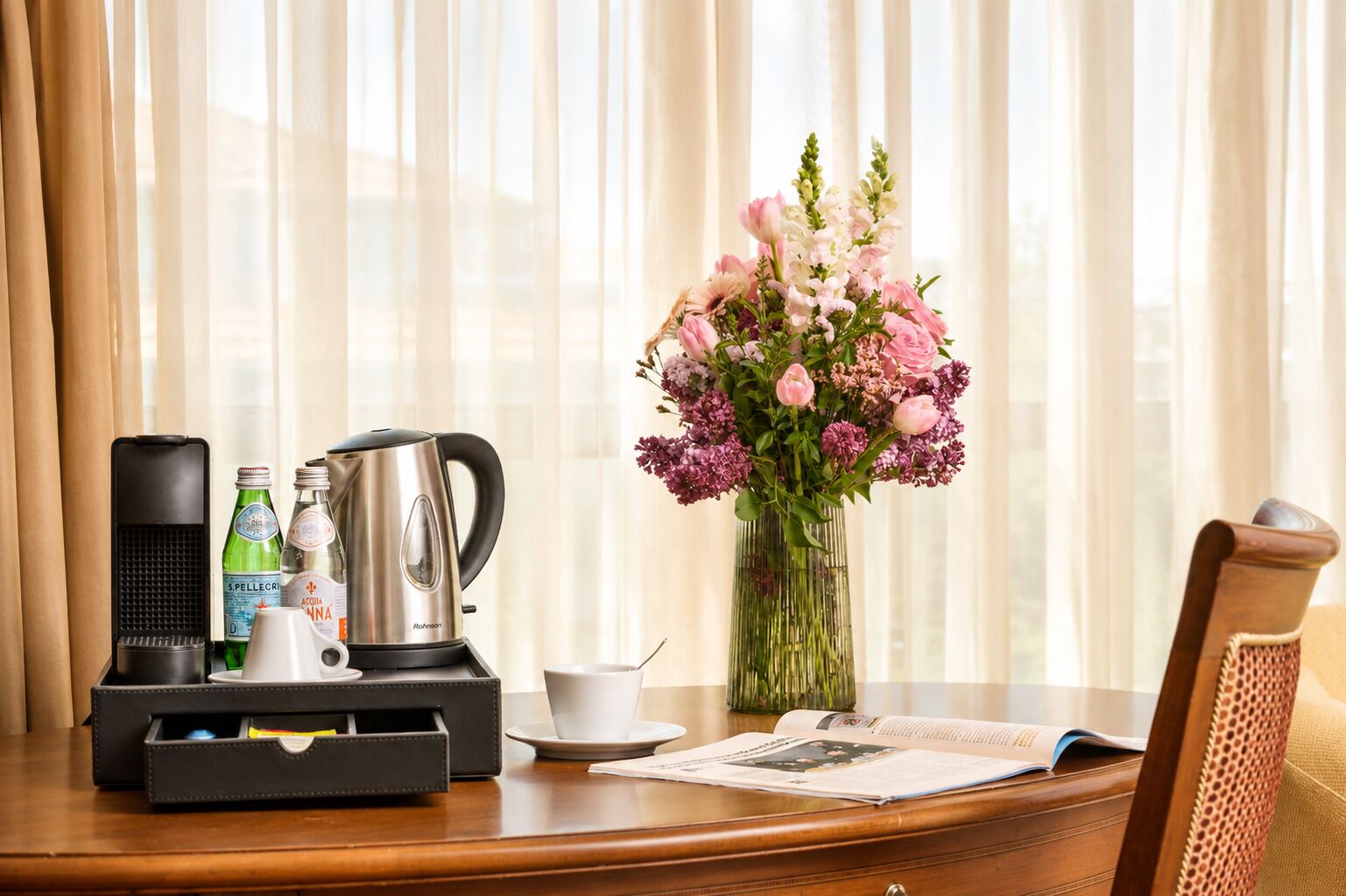 Une chambre d’hôtel équipée d’une machine à café, d’un service à thé et d’un vase de fleurs posé sur une table en bois.