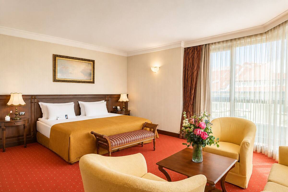 A hotel room with a bed, two bedside tables, a bench, two armchairs, a coffee table, and a window with curtains.