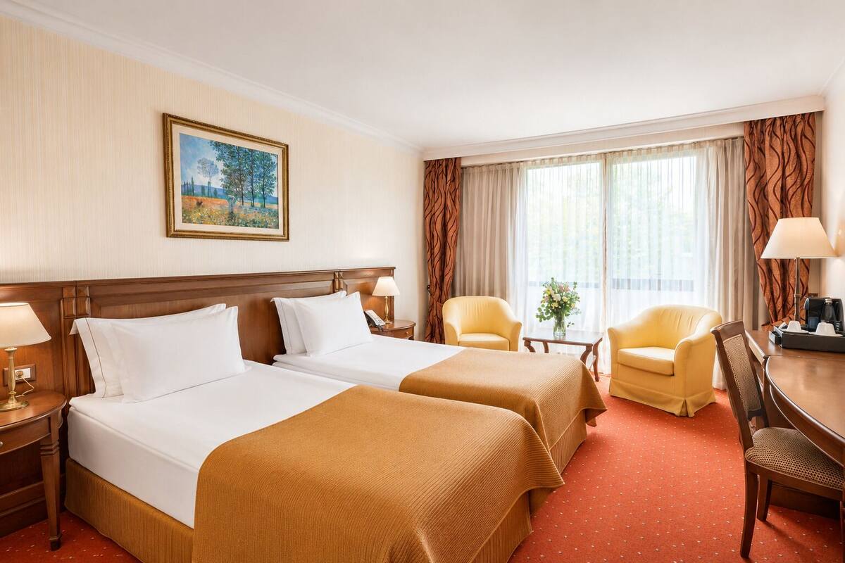 A hotel room with two beds, a desk, two chairs, a painting on the wall, and a window with curtains.