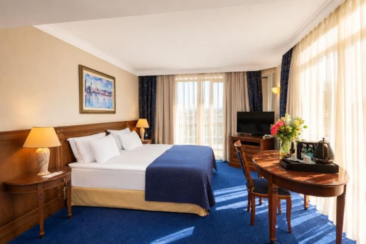 Quarto de hotel com cama, mesas de cabeceira, escrivaninha com cadeira, televisão e janela com cortinas.