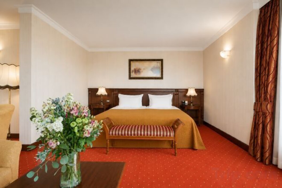 A hotel room with a bed, a bench, a vase with flowers, and a painting on the wall.