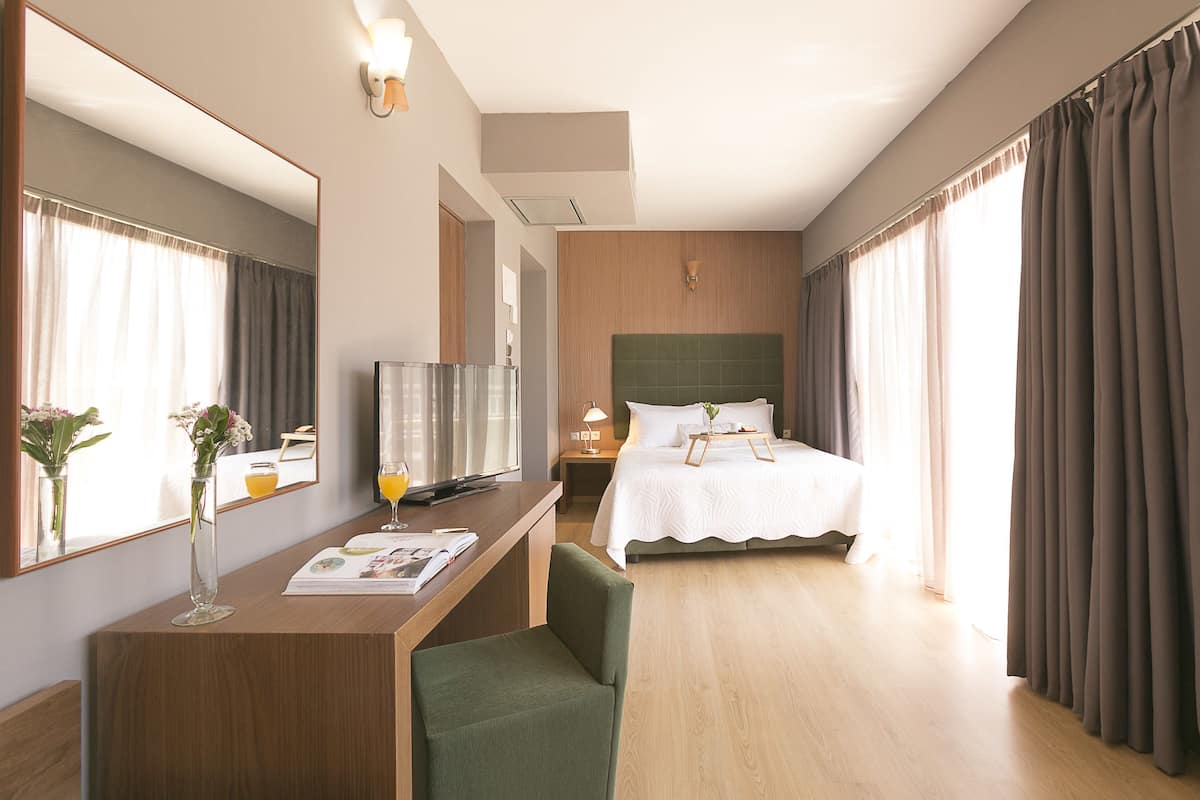 A hotel room with a bed, a desk with a chair, a TV, a bedside table with a lamp, and a large mirror.