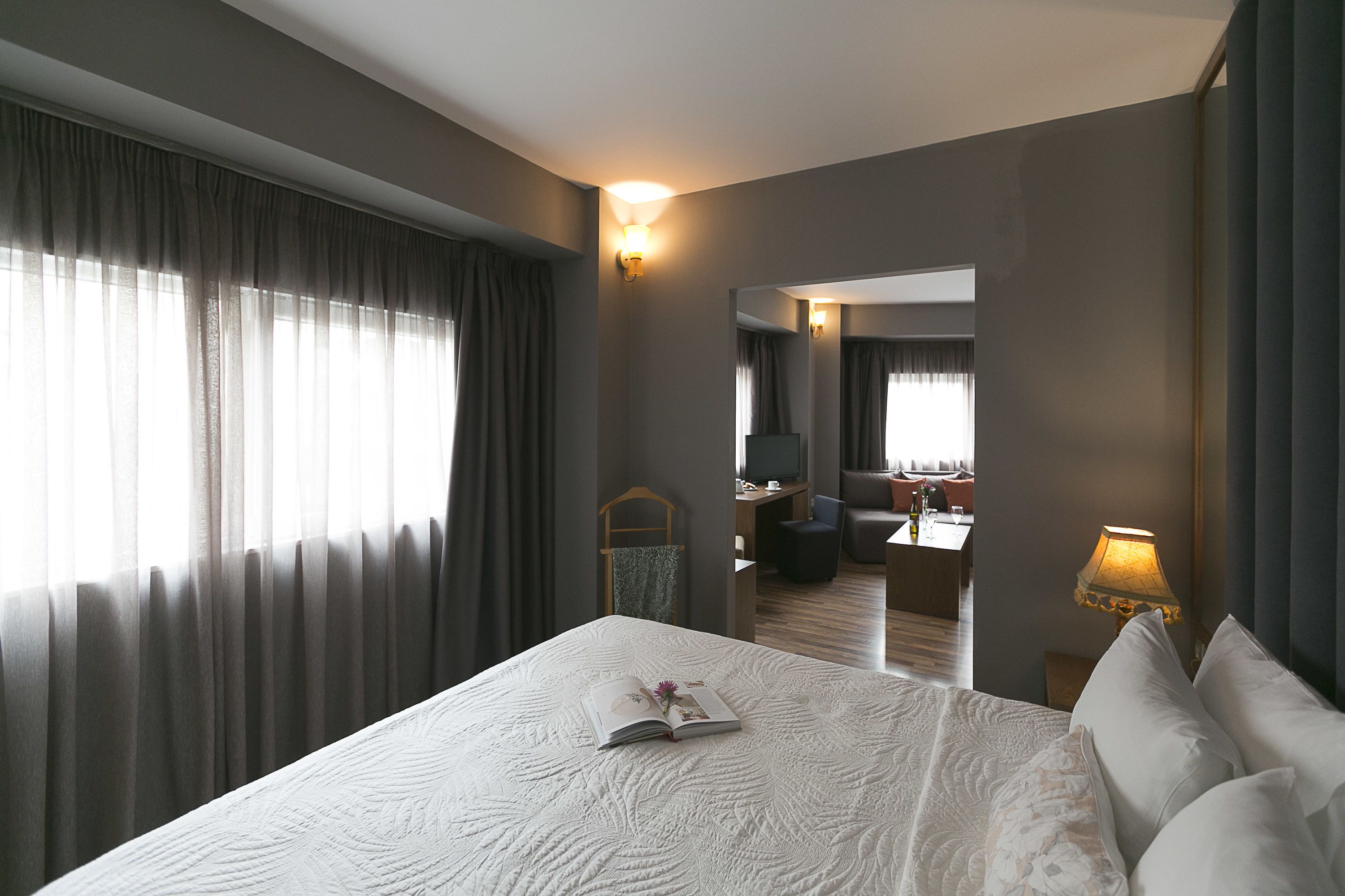 A hotel room with a large bed, a bedside table, a lamp, a TV, and a view of the hotel lobby.