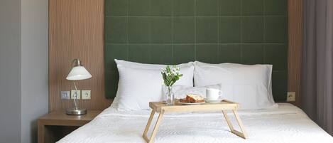 A neatly made bed with white linens, a bedside table with a lamp, a tray with bread, a cup, and a vase with flowers.