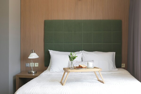 Una cama bien tendida con sábanas blancas, una mesita de noche con una lámpara, una bandeja con pan, una taza y un florero con flores.