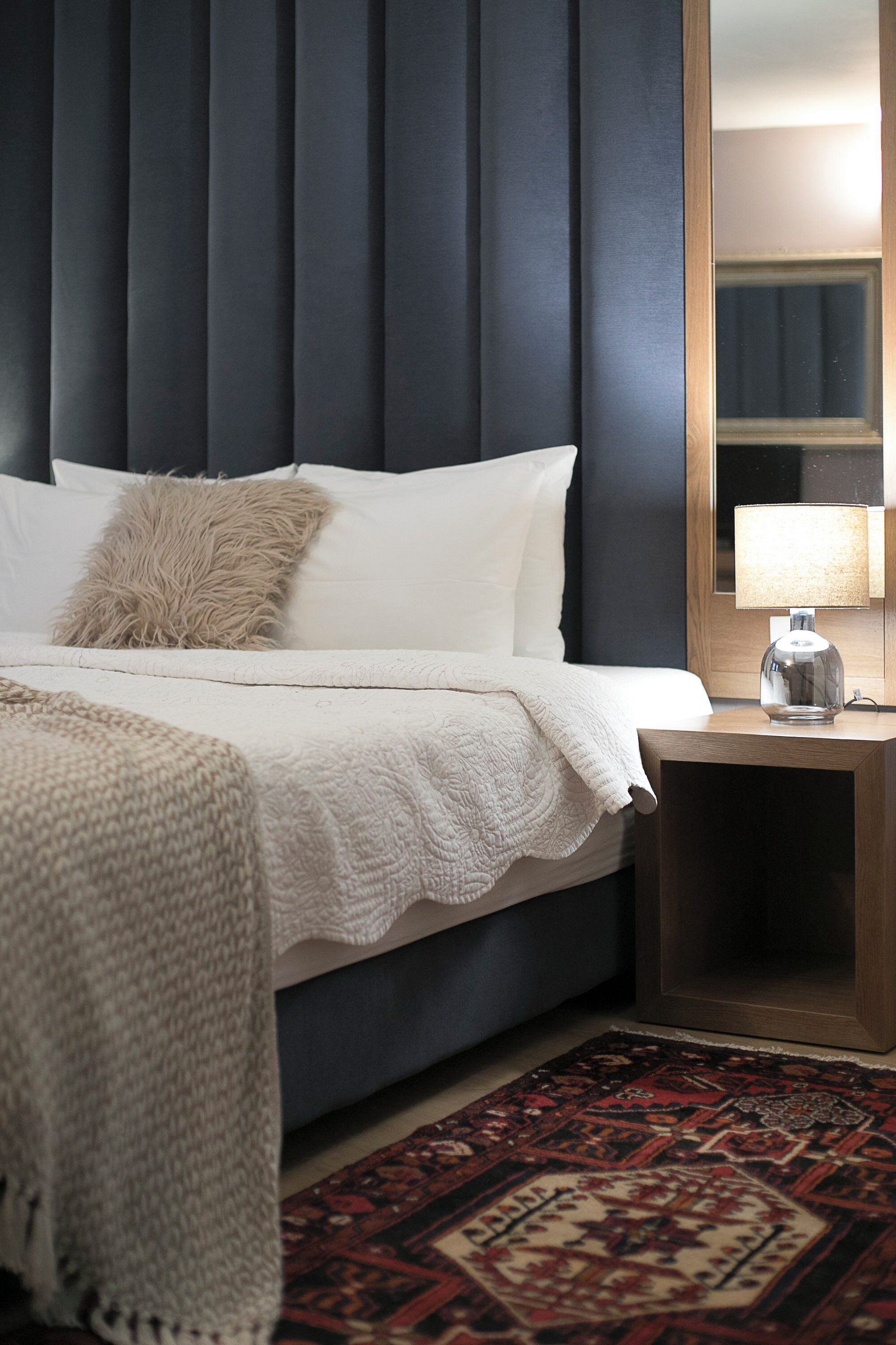 A neatly made bed with a white comforter and a fluffy pillow, a wooden nightstand with a lamp, and a patterned rug on the floor.