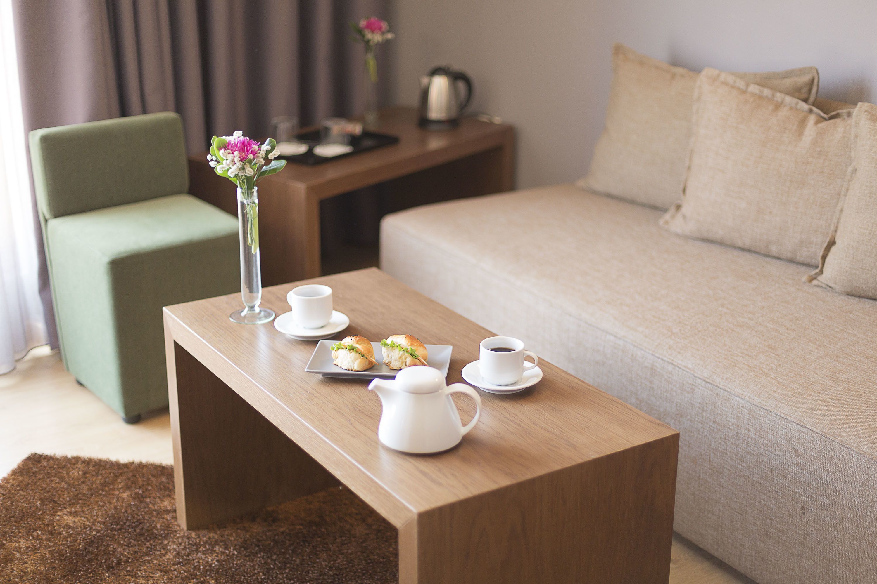 A neatly arranged room with a wooden coffee table, a green armchair, a beige sofa, and a small side table with a vase of flowers and a kettle.