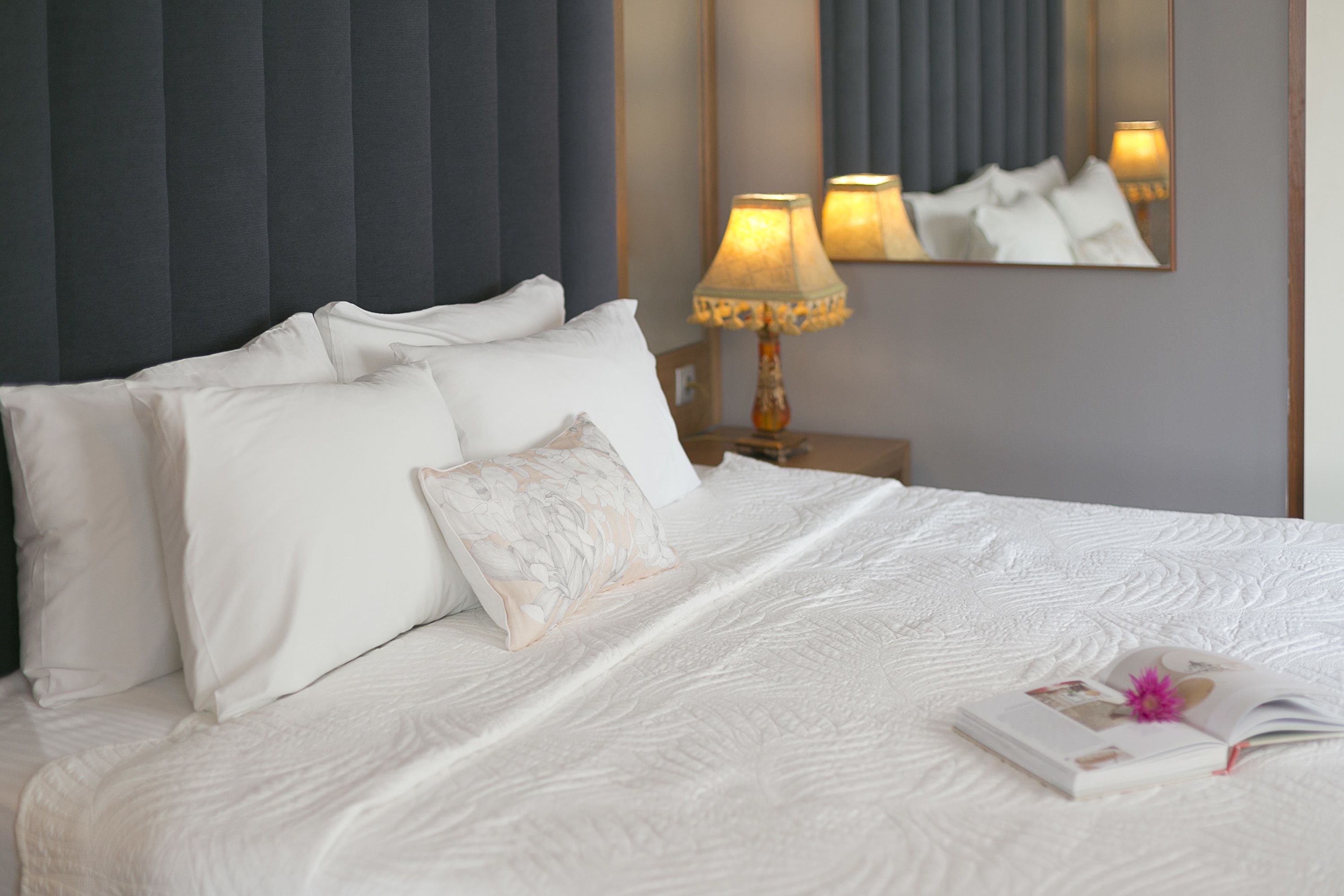 A neatly made bed with white linens and a floral pillow, a bedside table with a lamp, and a mirror reflecting the bed and lamp.