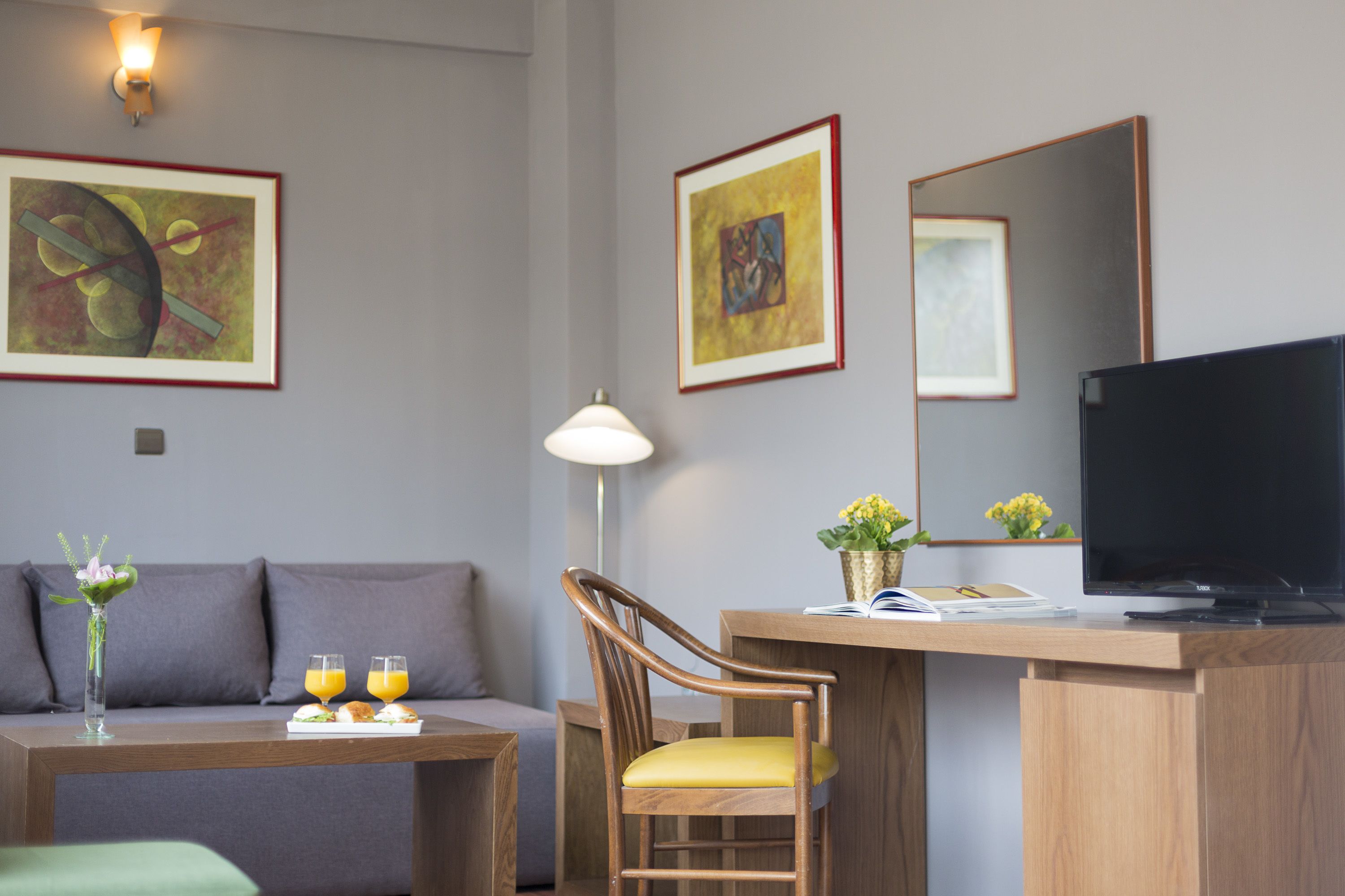 Quarto de hotel com sofá, escrivaninha com computador, cadeira, luminária e obras de arte nas paredes.