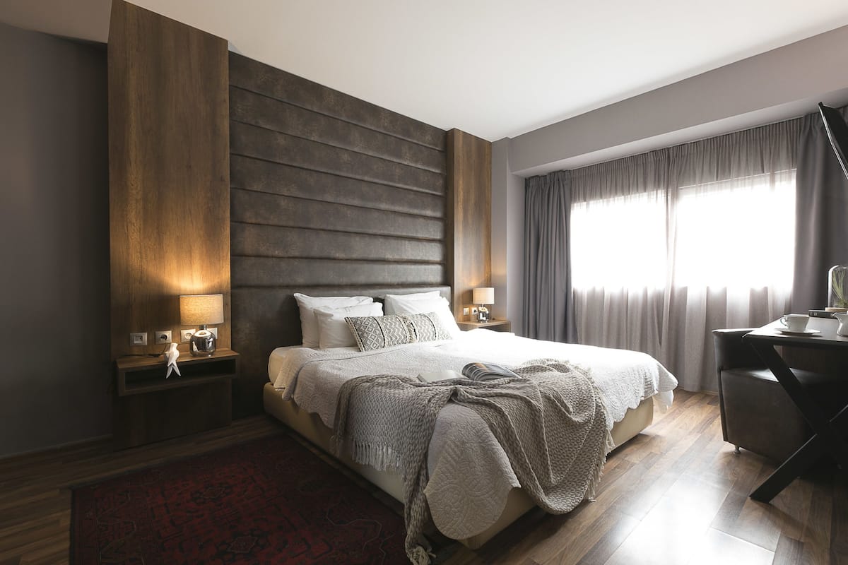 A modern hotel room with a large bed, bedside tables with lamps, a desk, and a view of the outdoors through large windows.