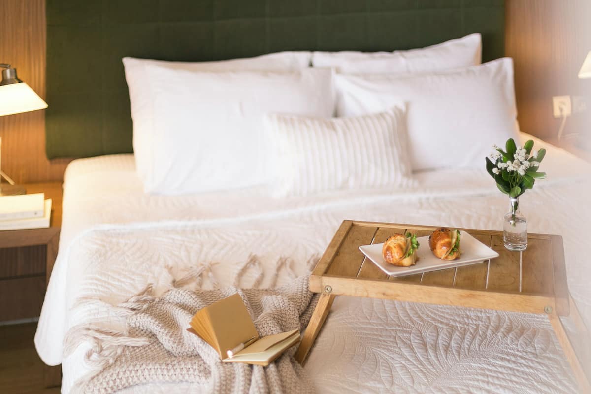 A neatly made bed with white linens, a tray with croissants, a book, and a small vase with flowers on a wooden tray.