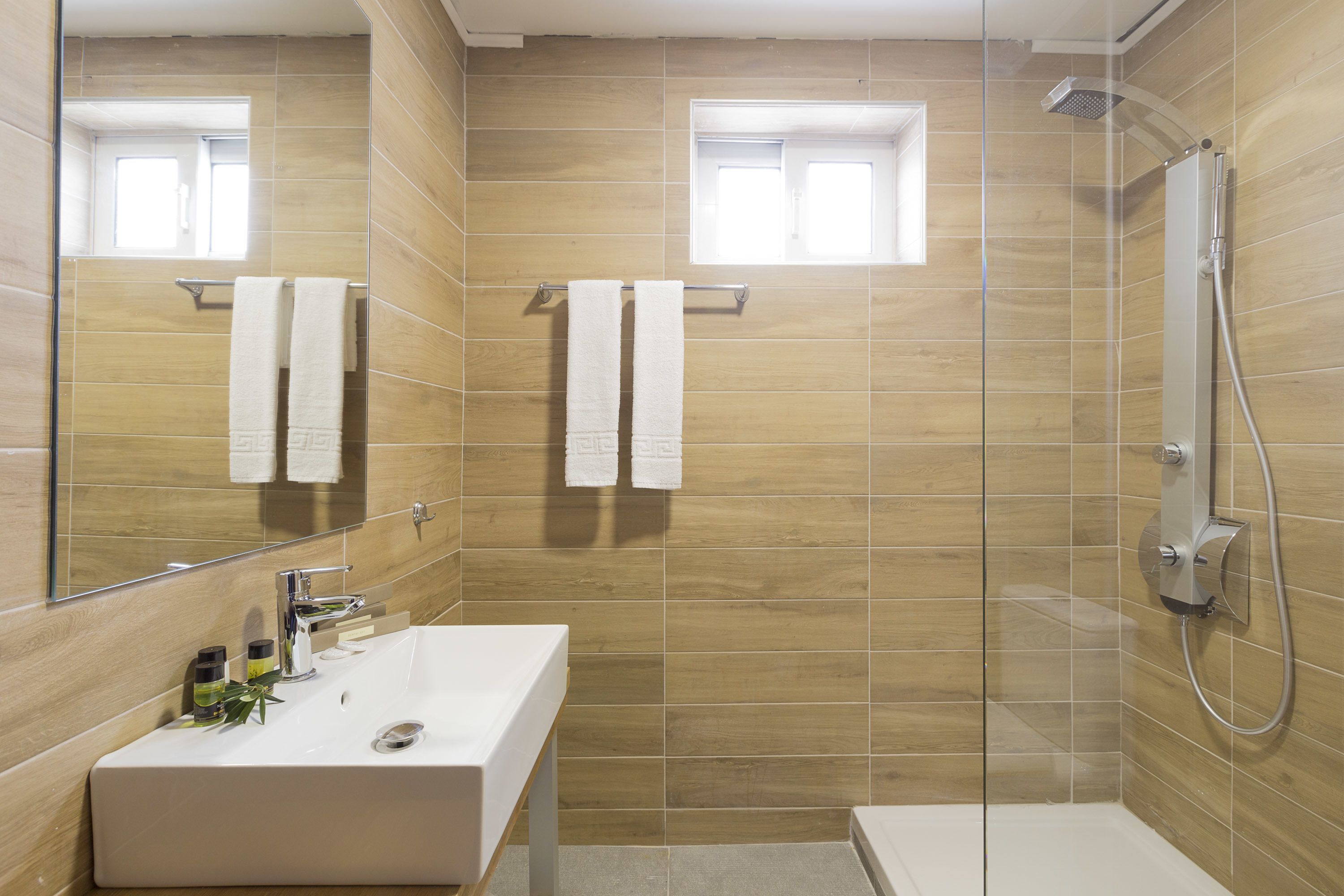A modern bathroom with a large mirror, a rectangular sink, a shower with a handheld showerhead, and two towels hanging on a rack.