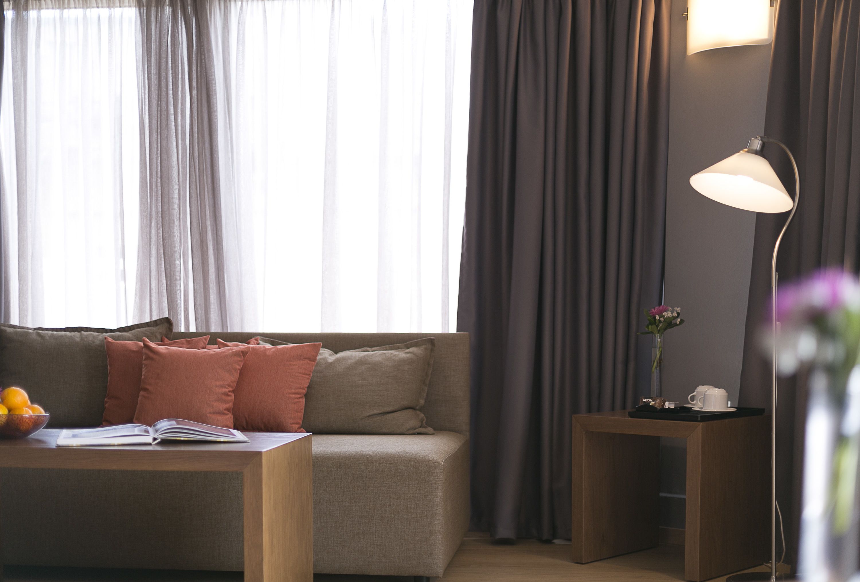 Una sala de estar moderna con un sofá, una mesa de centro con un libro y un cuenco de fruta, una mesita auxiliar con una lámpara, y un jarrón con flores.