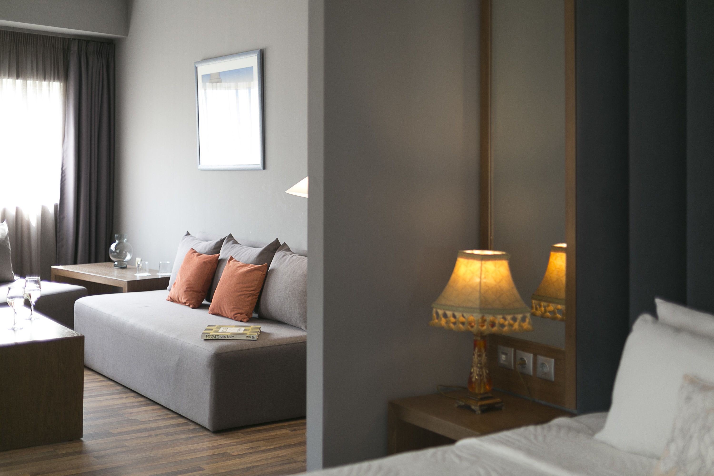 Una habitación de hotel con sofá, mesita de noche con lámpara, cama y ventana con cortinas.