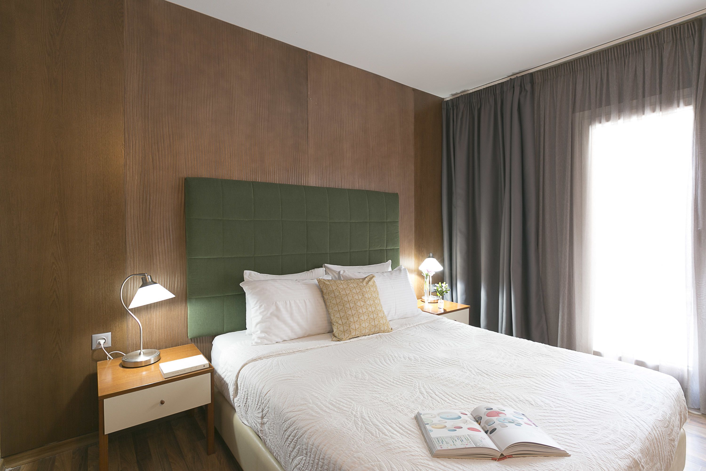 A neatly made bed with a green headboard, two bedside lamps, and a book on the bed.
