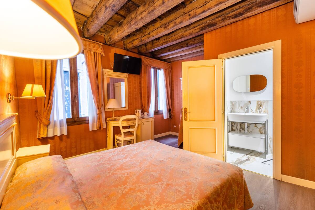 A room with a bed, a desk, a chair, a television, a mirror, and a bathroom with a sink and mirror.
