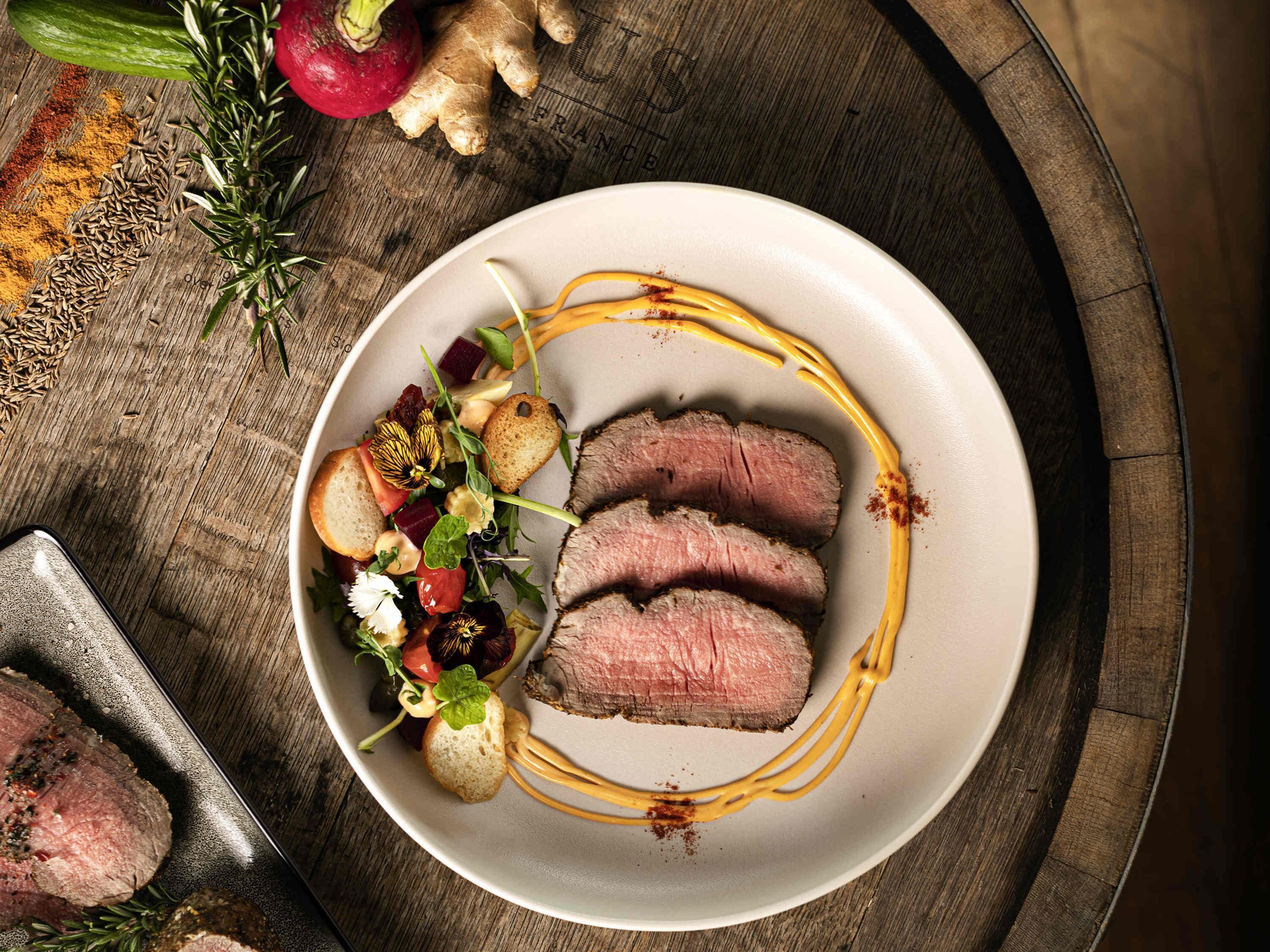 A plate of sliced meat with a side salad and bread on a wooden table.