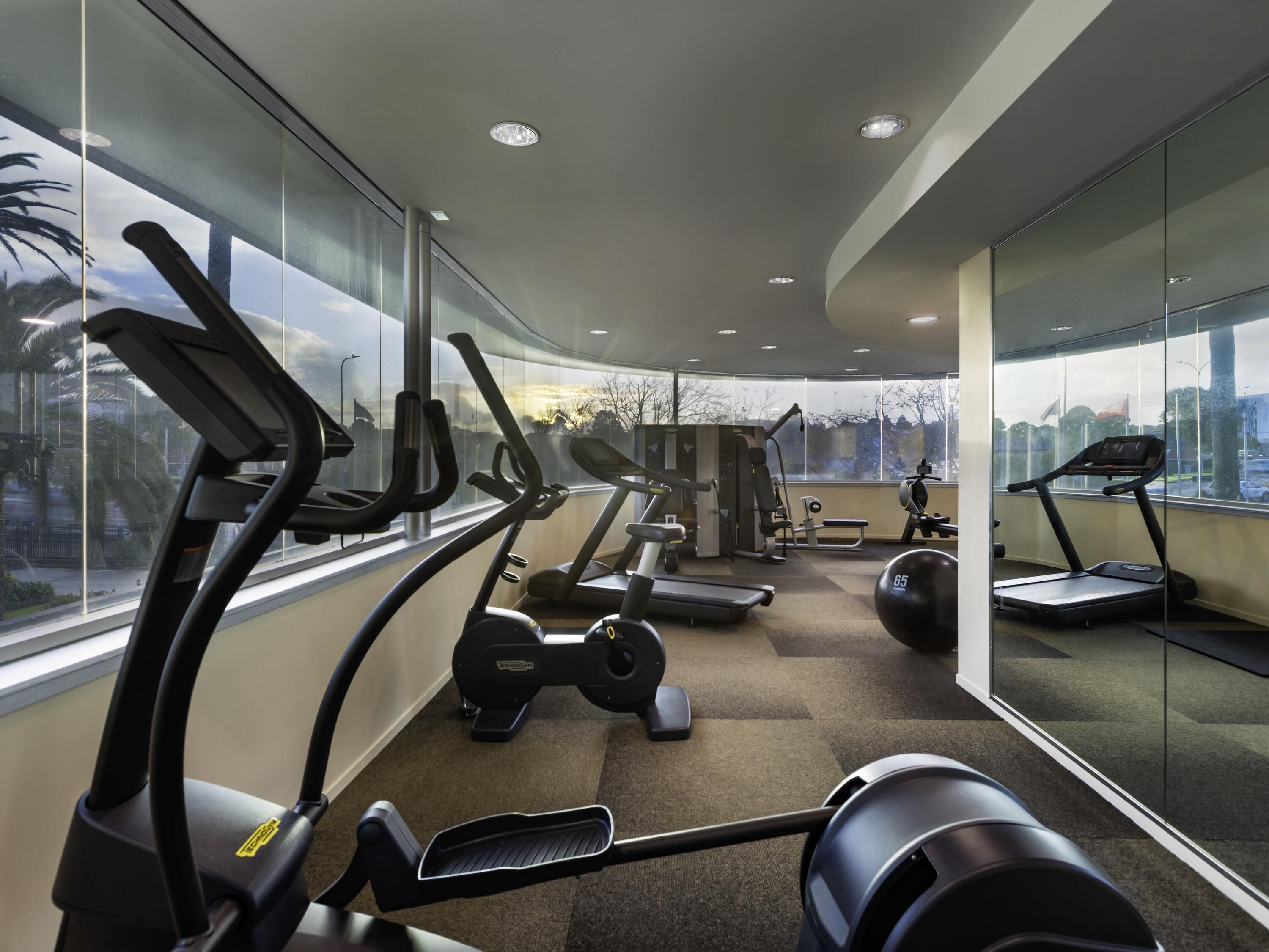 A modern gym with cardio machines, weight stations, and a view through large windows.