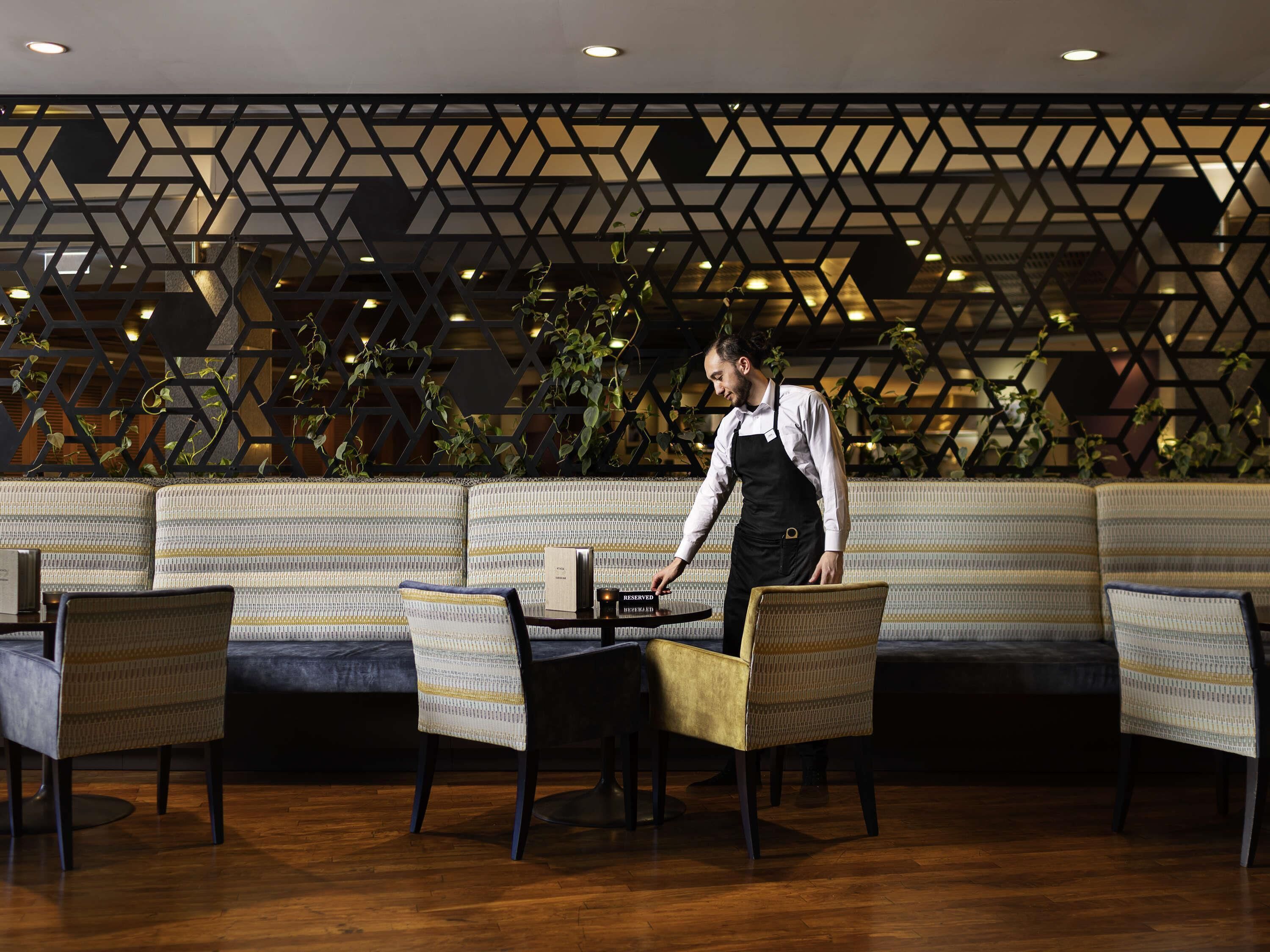 Un restaurante con un diseño moderno de pared geométrica, asientos acolchados y un camarero atendiendo una mesa.